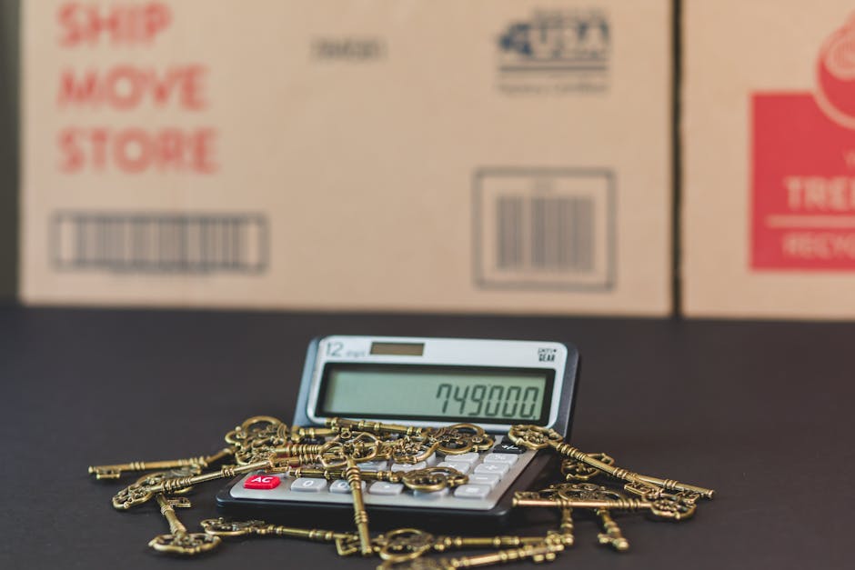 A calculator displaying the number 749800 sits on a flat surface, with several gold-colored keys and a gold-colored keychain placed atop it. Behind the calculator are two large cardboard boxes with printed labels, including barcodes and handling instructions such as 'Ship, Move, Store' and 'Fragile,' indicating ongoing packing or relocation activities. The scene appears to be inside a property or storage area, with the boxes positioned near an open doorway or loading zone, suggesting a home relocation or moving process. The lighting in the environment is neutral, focusing on the objects in the foreground, which relate to the packing and moving services offered by Man and Van Cranford, a company specializing in removals. The arrangement highlights the logistics of packing and furniture transport involved in house removals, as well as the detailed handling of items during a home move.