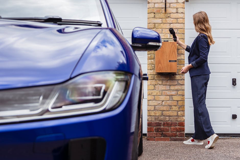 A woman with shoulder-length hair, dressed in a navy blazer and striped trousers, is standing beside a brick wall and white garage door, attaching a black strap to secure a wooden furniture piece inside a blue van during a home relocation process. The van, positioned on a driveway, is partially visible in the foreground with the front headlight and side mirror showing. She is wearing white sneakers with red accents and appears to be preparing the furniture for transport as part of a professional removals service by Man and Van Cranford. The scene captures the loading process, with attention to securing household items with packing materials like fabric and straps, supporting efficient furniture transport and moving logistics in the context of a house removal.