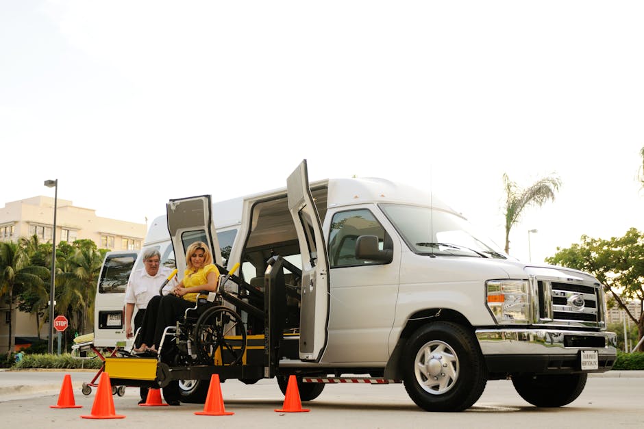 A white wheelchair-accessible van with its side door open, positioned on a paved street in a suburban area with palm trees and modern buildings in the background. Two individuals are present; one elderly man dressed in a white shirt and glasses, standing beside the vehicle, and a woman in a yellow top seated in a wheelchair, positioned near the edge of the van’s interior. The wheelchair is equipped with yellow safety features, and orange traffic cones are arranged around the van to delineate the loading area. The scene is outdoors during daytime with bright, natural lighting, illustrating a home relocation process involving furniture transport and packing materials, such as the wheelchair and safety cones, which support the context of moving services by Man and Van Cranford.