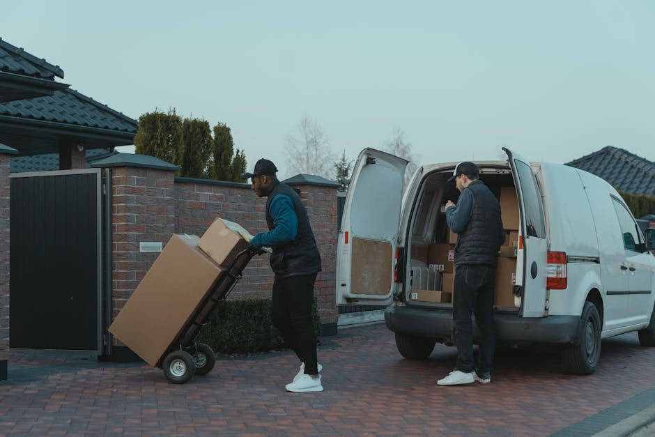 Two movers from Man and Van Cranford are engaged in a home relocation process, loading and unloading cardboard boxes and furniture in a quiet residential neighbourhood. One mover, dressed in a dark vest and cap, is pushing a two-wheeled trolley with large cardboard boxes towards the rear of a white van parked on a paved driveway. The van's hatchback doors are open, revealing additional packed boxes inside, with some wrapped in plastic or placed in cardboard cartons. The second mover, wearing a black cap, gloves, and a dark jacket, is inside the van, organizing or retrieving boxes for further transport. The environment suggests a peaceful street with houses featuring dark tiled roofs, tidy brick walls, and hedges in the background. The scene captures the logistical steps involved in packaging, carrying, and loading belongings during a professional house removal, with equipment such as trolleys and blankets visible to facilitate safe and efficient furniture transport, aligning with the services offered by Man and Van Cranford for residential moves and removals.