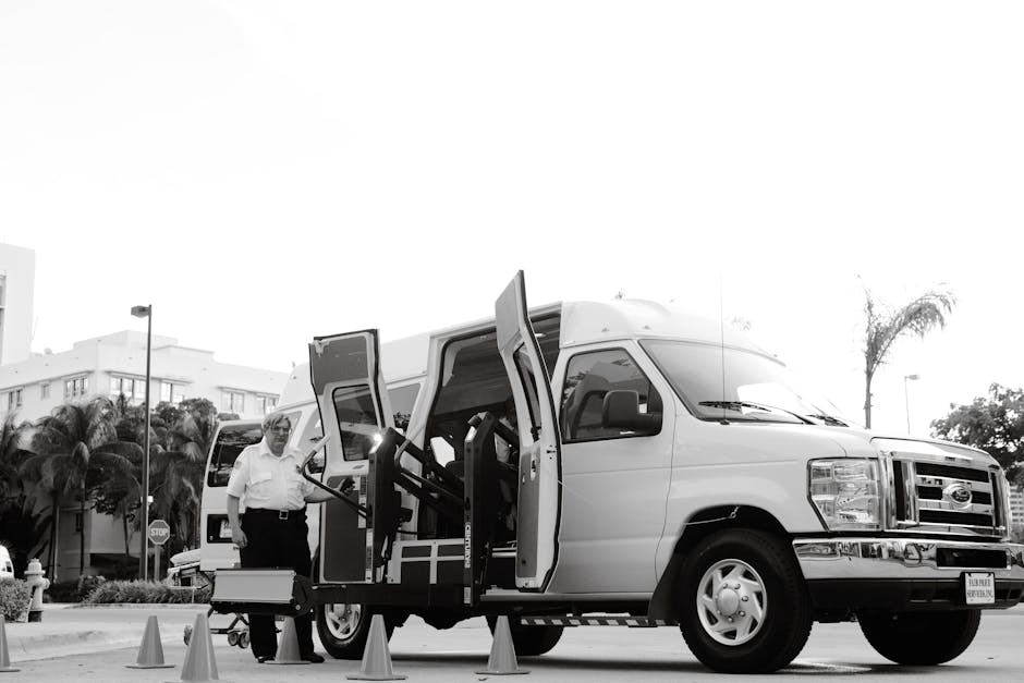 A white wheelchair-accessible van with its side door open, positioned on a paved street in a suburban area with palm trees and modern buildings in the background. Two individuals are present; one elderly man dressed in a white shirt and glasses, standing beside the vehicle, and a woman in a yellow top seated in a wheelchair, positioned near the edge of the van’s interior. The wheelchair is equipped with yellow safety features, and orange traffic cones are arranged around the van to delineate the loading area. The scene is outdoors during daytime with bright, natural lighting, illustrating a home relocation process involving furniture transport and packing materials, such as the wheelchair and safety cones, which support the context of moving services by Man and Van Cranford.