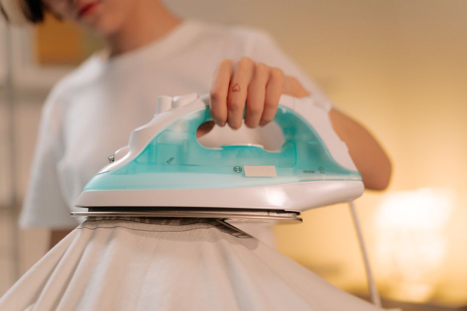A person in a white shirt is shown pressing an iron onto a piece of fabric, likely a bedsheet or clothing, with the iron positioned horizontally on top of the fabric. The background is blurred but suggests an indoor setting, possibly a home or an apartment with warm lighting. The individual is holding the iron's handle with their right hand, and only part of their face and arm are visible. The scene relates to household chores such as packing or preparing items for a home relocation, integrated into the context of house removals and moving services. This visual depiction aligns with the theme of packing, ironing, and preparing belongings during furniture transport or home moves. Man and Van Cranford offers professional removals, ensuring careful handling of all household items during the loading process.