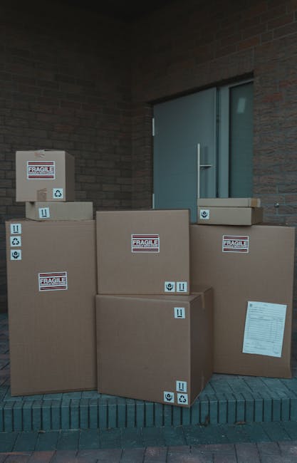 Several large cardboard boxes stacked outside a building, positioned on a brick-paved area near a blue door. The boxes are secured with packing tape and feature labels indicating fragility, recycling