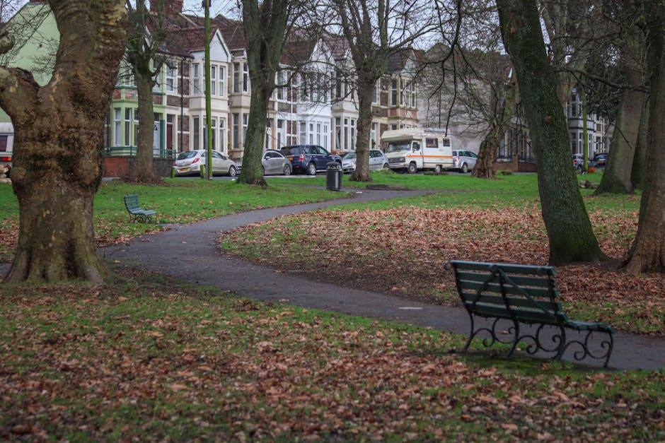 A paved pathway meanders through a park area surrounded by large, mature trees with thick trunks and sprawling branches. Fallen leaves cover the ground, indicating an autumn setting. In the foreground, a green metal bench with decorative scrollwork is positioned on the right side, facing the pathway. Further along the path, another bench can be seen on the left side, also made of metal with a similar design. In the background, residential buildings with bay windows and multi-storey façades line the street, with parked cars visible along the curb. A white van with an orange stripe, likely part of a house removals or moving service, is parked close to the sidewalk near the buildings. The scene suggests an urban park adjacent to residential properties, with [COMPANY_NAME], such as Man and Van Cranford, potentially involved in household relocation or furniture transport activities nearby during a home moving process.