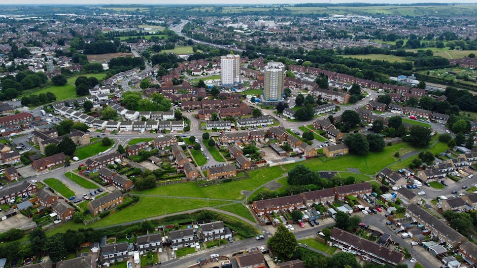 An aerial view of a residential neighbourhood showing a mix of detached and semi-detached houses with small gardens and driveways. The houses are situated along a tree-lined street with parked cars and moving vehicles visible. To the left, there is a large green park area with dense trees, lawn, and garden plots, indicating a peaceful community environment. The scene is captured in bright daylight with natural sunlight enhancing the vivid greens of the trees and grass. This image provides visual context for house removals and furniture transport services carried out by Man and Van Cranford, highlighting the typical suburban setting involved in local home relocation and packing and moving activities.