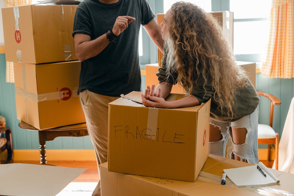 A smiling man with a beard and curly hair, wearing a teal T-shirt and grey trousers, stands inside a bright room with large, arched windows in the background. He is holding a medium-sized cardboard bo