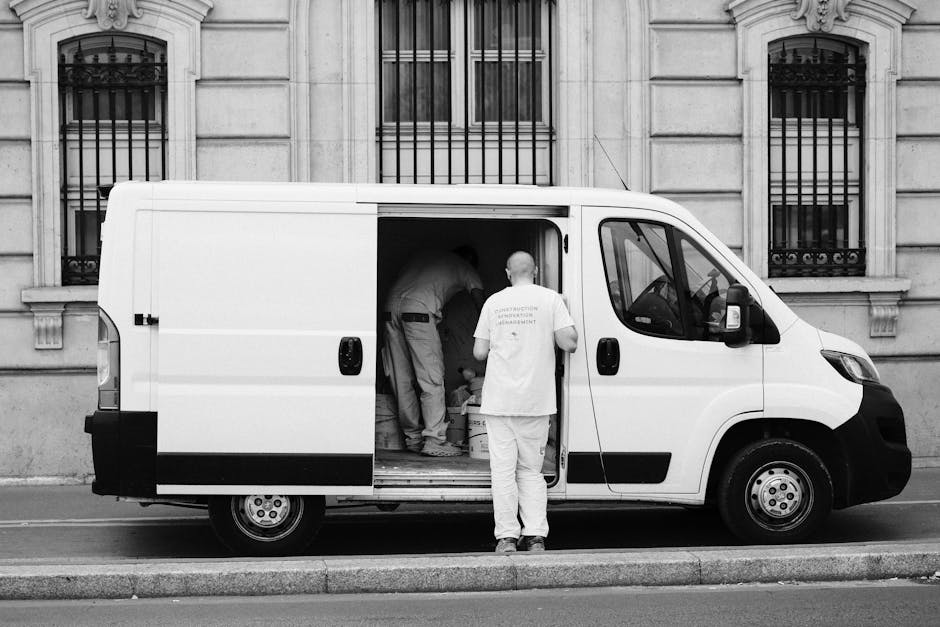 A white commercial van with its sliding side door open is parked on a city street in front of an ornate stone building with decorative window bars. Inside the van, several movers are actively engaged in loading boxes, furniture, and packing materials, including cardboard boxes, plastic-wrapped items, and wooden furniture, onto the vehicle. One mover, dressed in a white t-shirt and light trousers, stands outside the van facing inward, supervising or preparing to assist with the packing process. The workers appear focused on their task, with some bent over placing items inside the van. The environment suggests a home relocation or furniture transport scene, with important moving equipment such as trolleys and straps possibly used but not visible. The street’s curb and pavement are visible, indicating the vehicle is parked in compliance with local unloading regulations often relevant for Cranford removals, as addressed on the company’s webpage about Heathrow unloading rules for removal vans. This scene captures the logistical effort involved in packing and loading household or office belongings for transportation during a house move.