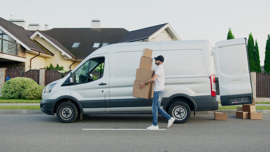 A man wearing a white t-shirt and jeans, and a woman in a black top, denim skirt, and white cap, stand beside the open rear doors of a white moving van parked on the pavement under a partly cloudy sky. Inside the van, numerous brown cardboard boxes of varying sizes are neatly stacked, some secured with plastic wrap, ready for the packing and moving process involved in home relocation. The woman appears to be holding a clipboard or list, likely coordinating the loading or unloading of furniture and packing materials, while the man is checking his phone or a device. The scene depicts the loading process typical during furniture transport or house removals, with the van positioned near an entrance to a property or street, supported by the services of Man and Van Cranford. This image exemplifies the logistical steps in packing, organizing, and transporting household belongings as part of professional removals service in Cranford, referencing Heathrow unloading rules for removal vans with clean, natural lighting highlighting the environment.