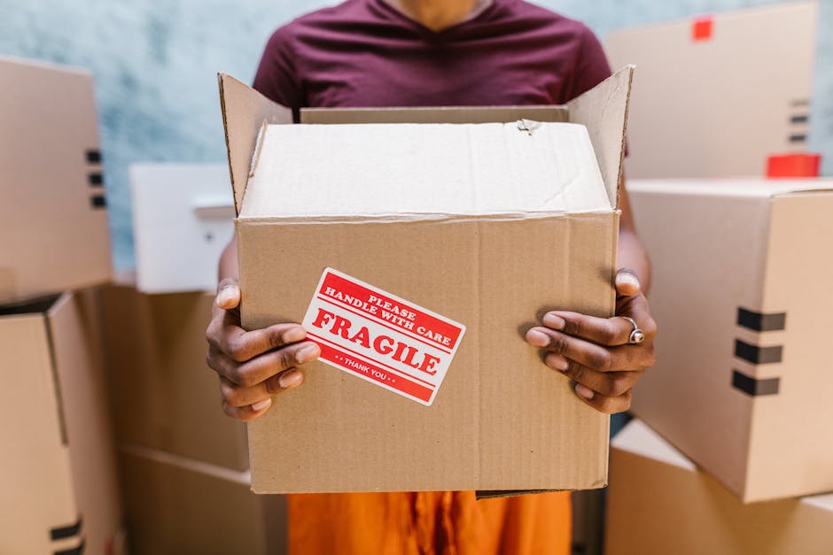 A person holding a medium-sized cardboard box with both hands inside a room filled with stacked similar boxes. The box has a red and white 'Fragile Handle with Care' sticker on the front. The individual is wearing a maroon top, and their hands are visible, showing short nails and a silver ring on one finger. In the background, more cardboard boxes are arranged on the floor and against the wall, some with black tape sealing the lids. The setting appears to be a home or warehouse environment, indicating a packing or home relocation process. The lighting is bright, highlighting the labels and packing materials, which include cardboard and plastic wrapping, typical for furniture transport and moving services. Man and Van Cranford occasionally manages packing and moving tasks, emphasizing careful handling of fragile items during residential removals.