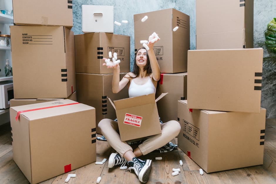 A young woman with long brown hair, dressed in a white tank top and beige trousers, is sitting cross-legged on a wooden floor inside a room, surrounded by numerous cardboard moving boxes. She is smiling and throwing small white packing peanuts into the air, creating a playful scene. Several of the boxes, some sealed with red tape and others with black printed labels, are stacked around her at different heights, some on the floor and others stacked against the wall. Behind her, a tall stack of boxes reaches up to her shoulders, with additional boxes placed on a side table to her left. The room's background features a textured light blue wall, a small white shelf with a few decorative items, and a green leafy plant on the far right. The lighting appears natural, illuminating the scene evenly. This image reflects home relocation, packing, and moving processes, with [COMPANY_NAME] offering professional removals services in the context of a house move to ensure smooth furniture transport and logistical organization, aligned with the page about London Borough of Hounslow move permit fines and removals.