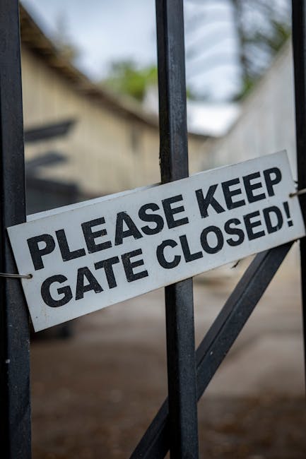 Close-up image of a metal gate with a white rectangular sign attached that reads 'PLEASE KEEP GATE CLOSED!' in bold black letters. The gate is part of a fenced area, with the background showing a blurred view of outdoor environment including some greenery, a wooden structure, and overcast sky. The sign is secured with wire, and the gate's vertical and diagonal bars are visible, suggesting it is used for property access control. This image exemplifies perimeter security during house removals or relocations, highlighting the importance of gate management in transport and moving processes. Natural lighting and a neutral colour palette support a professional, factual presentation relevant to home relocation logistics.