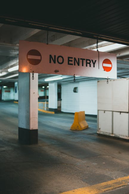 A large rectangular 'NO ENTRY' sign with black lettering and red circle symbols on a white background hangs from the ceiling of an underground parking garage, supported by metal chains. The parking area features concrete pillars, some painted with black and white stripes, and a yellow wheel stop block positioned on the asphalt floor. The space is dimly lit with artificial lighting, and in the background, there are white walls, parking bays, and indicating lines for vehicle placement. This restricted access zone may pose challenges during house removals and furniture transport, requiring careful planning when navigating through parking structures as part of home relocation services offered by Man and Van Cranford, especially when moving items along Bath Road, CRANFORD.