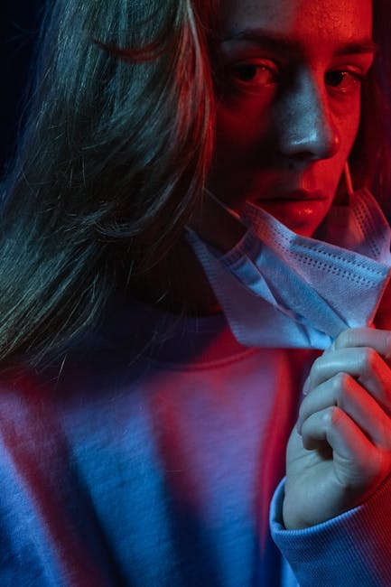 A woman wearing a light blue hoodie and a white disposable face mask is inside a property, likely preparing for a home relocation or moving process. Her long hair partially covers her face, which is partially visible as she looks slightly downward. She is holding the mask with her right hand, as if adjusting it, with the other hand not fully visible. The lighting in the room features a mix of blue and red tones, creating a moody atmosphere. The background suggests an indoor environment, possibly near the doorway or the entry point to a house, where packing and moving preparations are being made. Visible objects include the hoodie fabric, the mask, and an indoor setting indicating a staging area for furniture transport or packing for a move. This image, associated with [COMPANY_NAME], reflects the careful planning and precautions involved in house removals and furniture transport, supporting the theme of efficient and safe relocation services.