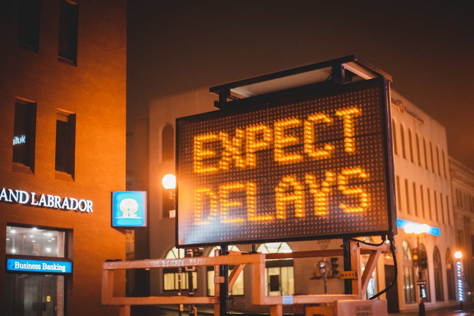 A large illuminated electronic signboard displaying the message 'EXPECT DELAYS' in bright orange lights, situated outdoors on a city street at night. The sign is mounted on a wheeled stand with visible support beams and electrical cables connected at the base. Surrounding the sign are nearby buildings with brick and white facades, illuminated by streetlights, including a building with the signage 'LAND & LABRADOR' and a visible blue-lit ATM or business entry. The scene captures a typical urban environment during early evening or night, with no visible moving equipment or furniture, emphasizing the importance of planning transportation logistics during house relocations, such as those offered by Man and Van Cranford, particularly around expected delays.