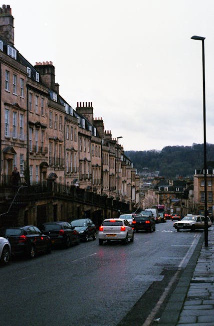 A street scene in Cranford featuring a row of classic sandstone terraced houses with chimney stacks and multiple stories, situated along the left side of the image. Parked cars line both sides of the street, with some vehicles in motion, indicating active street parking and traffic. The road is slightly wet, suggesting recent rain, and the sky is overcast, contributing to diffused natural lighting. To the right, a tall street lamp stands on the sidewalk, which is paved with rectangular stones. In the background, a hilly landscape with sparse tree coverage can be seen beyond the buildings. This setting, captured during daylight, is typical of residential areas where house removals and furniture transport activities may take place, with the scene providing context for urban moving logistics and packing procedures in Cranford, as exemplified by [COMPANY_NAME] in their house removal services.