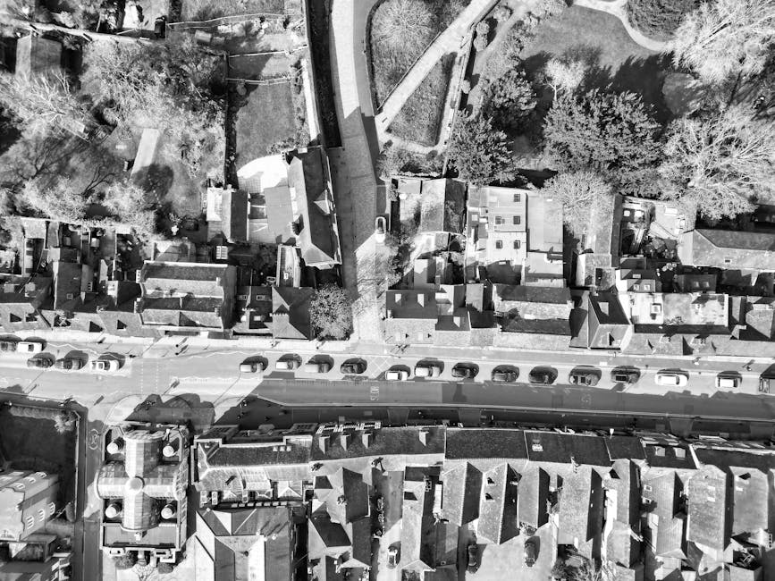 An aerial black-and-white photograph of a residential street showing parked cars lining both sides of the road. Directly above the street, several houses with pitched roofs, chimneys, and small yards are visible, with some yards containing trees and shrubs. A narrow footpath runs parallel to the street, separated by a curb, leading to the entrances of the houses. In the upper part of the image, a driveway with a van parked on it is visible near a garden area. The scene appears to capture a quiet suburban neighbourhood during daylight, with shadows cast by the buildings and trees. This image is relevant to house removals or relocation services, illustrating the typical environment where furniture transport and packing processes, managed by companies like Man and Van Cranford, are carried out during a home relocation or moving day.