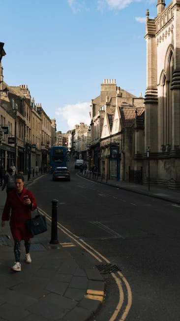 A street scene in Cranford featuring a row of classic sandstone terraced houses with chimney stacks and multiple stories, situated along the left side of the image. Parked cars line both sides of the street, with some vehicles in motion, indicating active street parking and traffic. The road is slightly wet, suggesting recent rain, and the sky is overcast, contributing to diffused natural lighting. To the right, a tall street lamp stands on the sidewalk, which is paved with rectangular stones. In the background, a hilly landscape with sparse tree coverage can be seen beyond the buildings. This setting, captured during daylight, is typical of residential areas where house removals and furniture transport activities may take place, with the scene providing context for urban moving logistics and packing procedures in Cranford, as exemplified by [COMPANY_NAME] in their house removal services.