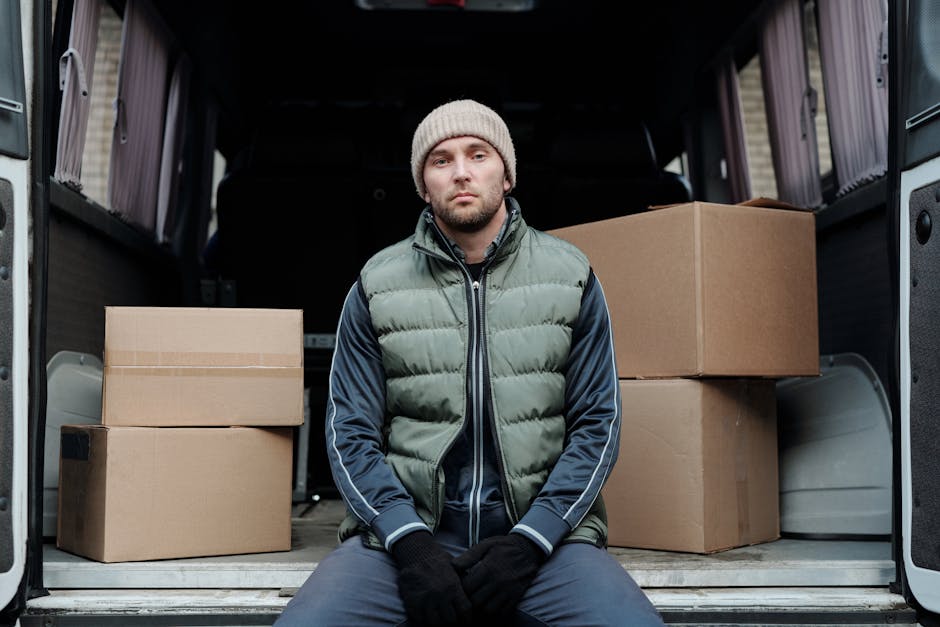A young man with a serious expression sits on the edge of an open van's loading area, surrounded by several large cardboard boxes of varying sizes, all securely closed. He is dressed in a grey beanie,