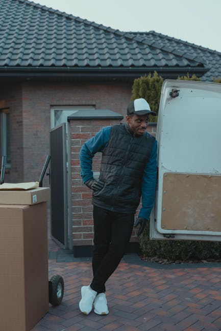 A young man with a serious expression sits on the edge of an open van's loading area, surrounded by several large cardboard boxes of varying sizes, all securely closed. He is dressed in a grey beanie,